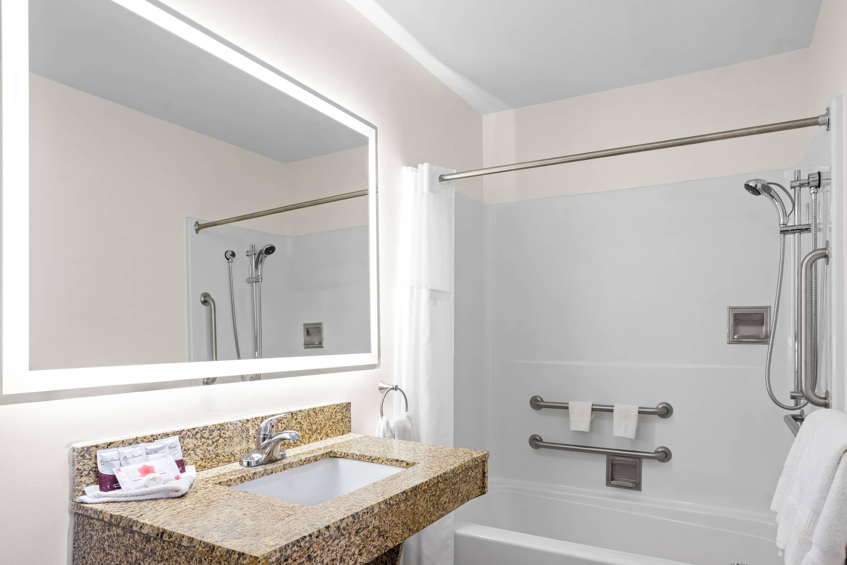 Un baño moderno con lavamanos de granito, un espejo grande, ducha a ras de suelo con barras de apoyo y bañera blanca.