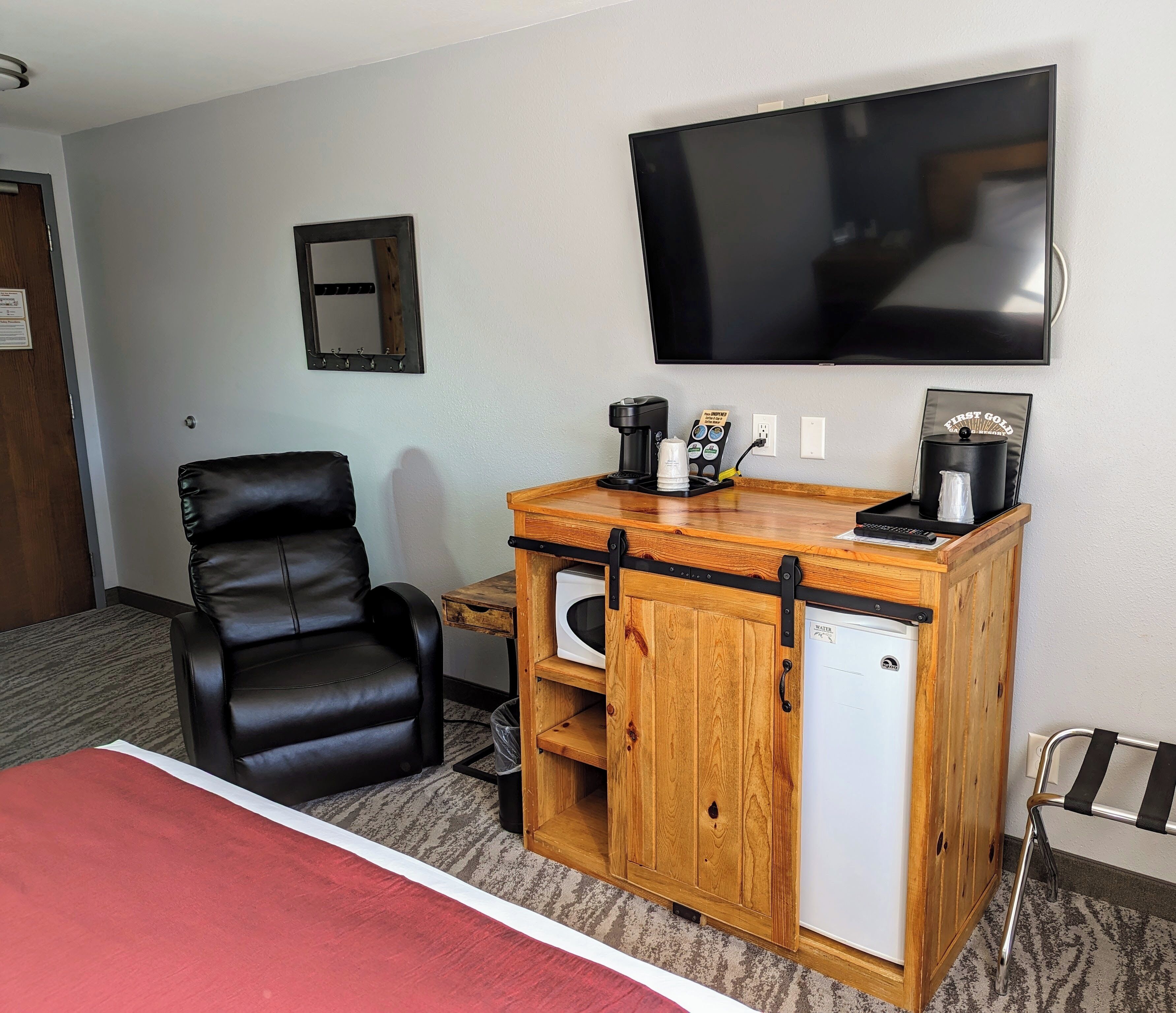 Habitación de hotel con una cama, un mueble de madera con cafetera, un televisor y un sillón de cuero negro.