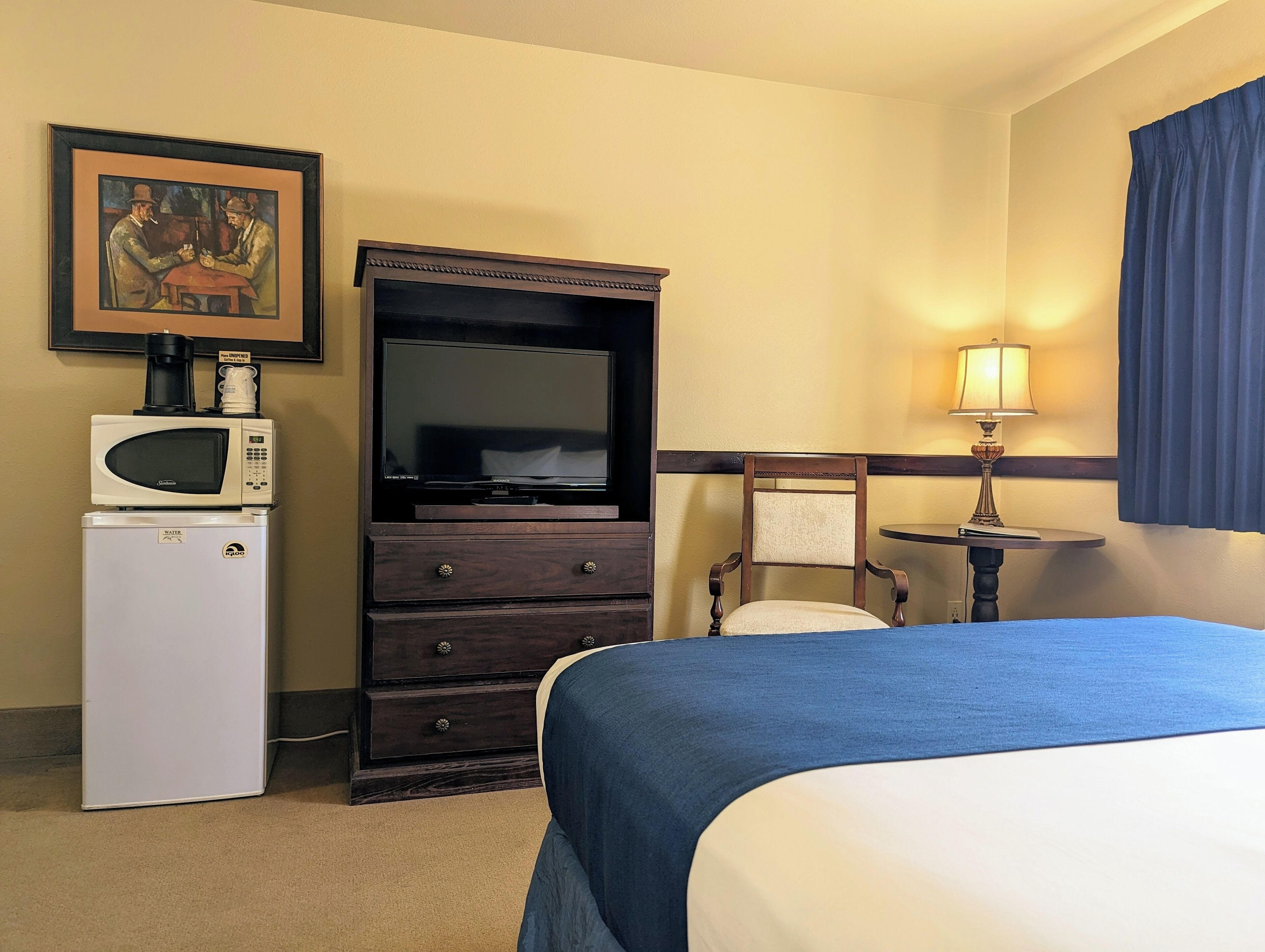 A hotel room with a bed, a TV, a microwave, a refrigerator, a chair, a table, and a painting on the wall.