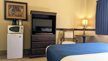 A hotel room with a bed, a TV, a microwave, a refrigerator, a chair, a table, and a painting on the wall.