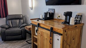 Ein kompakter Wohnbereich mit einem Ledersessel, einer kleinen Holzbar mit Kaffeemaschine und einem an der Wand montierten Fernseher.