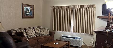 Una sala de estar con un sofá de cuero marrón, una mesa de centro de madera y una alfombra beige. Hay un cuadro en la pared y una ventana con cortinas.
