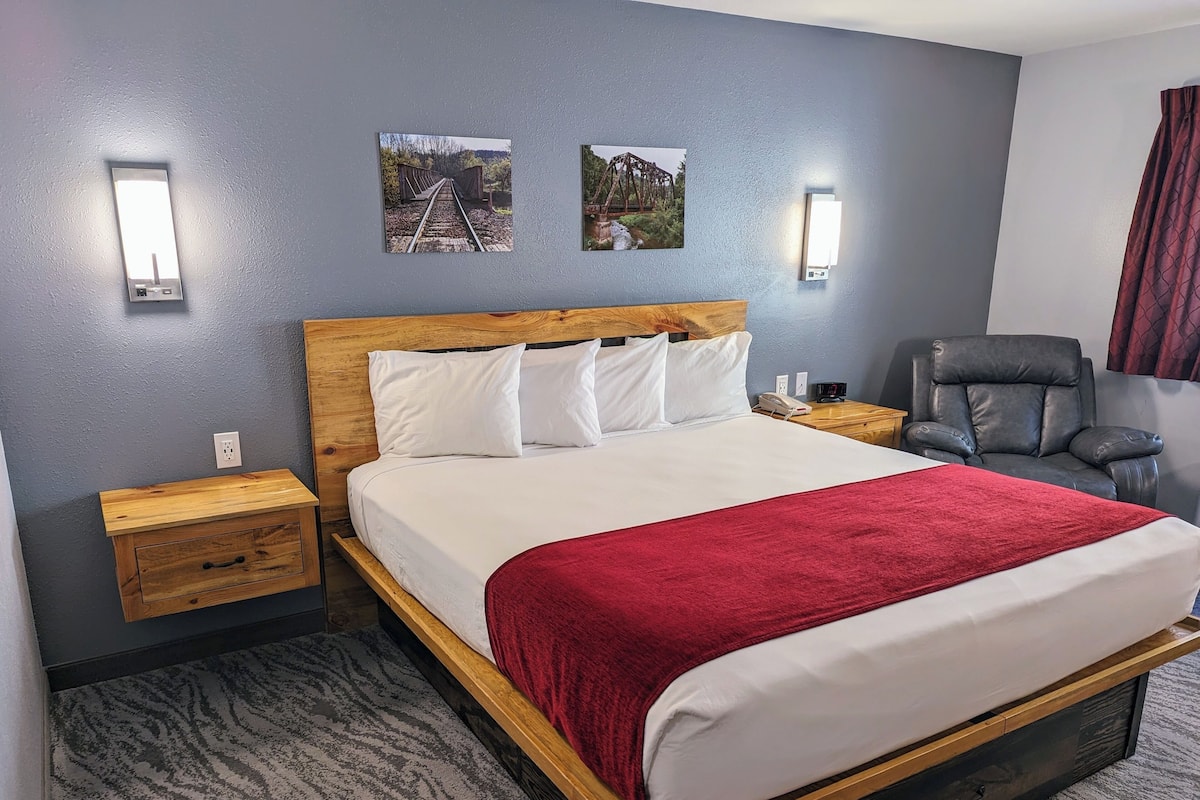 Une chambre d’hôtel avec un lit en bois, du linge de lit blanc, un plaid rouge, une table de chevet avec un téléphone, une chaise et deux lampes fixées au mur.