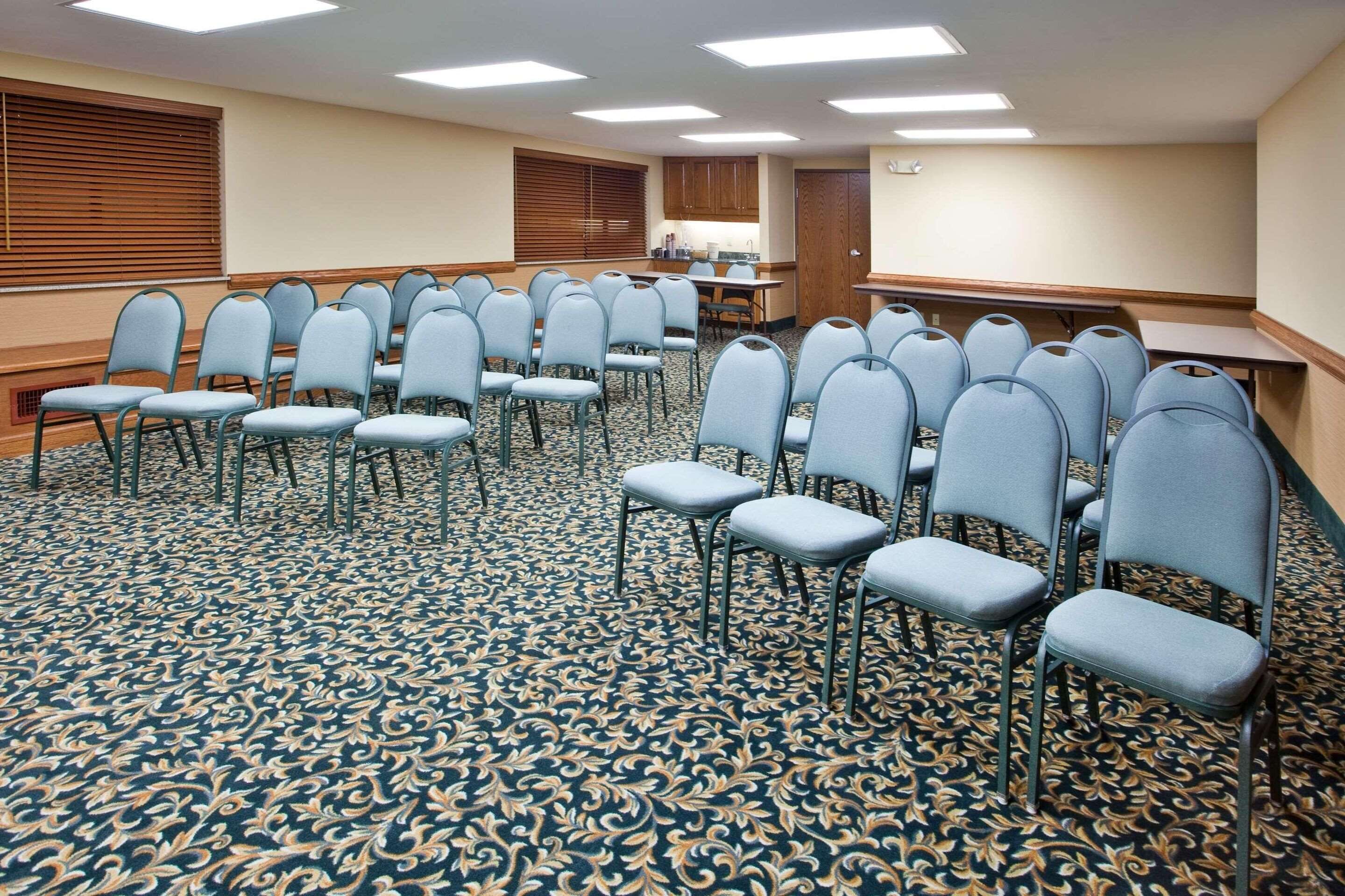 A conference room with rows of chairs, a kitchen area, and a large window with blinds.