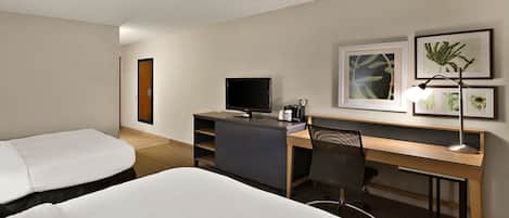 A hotel room with two beds, a desk with a computer, a chair, a TV, and framed botanical artwork on the wall.
