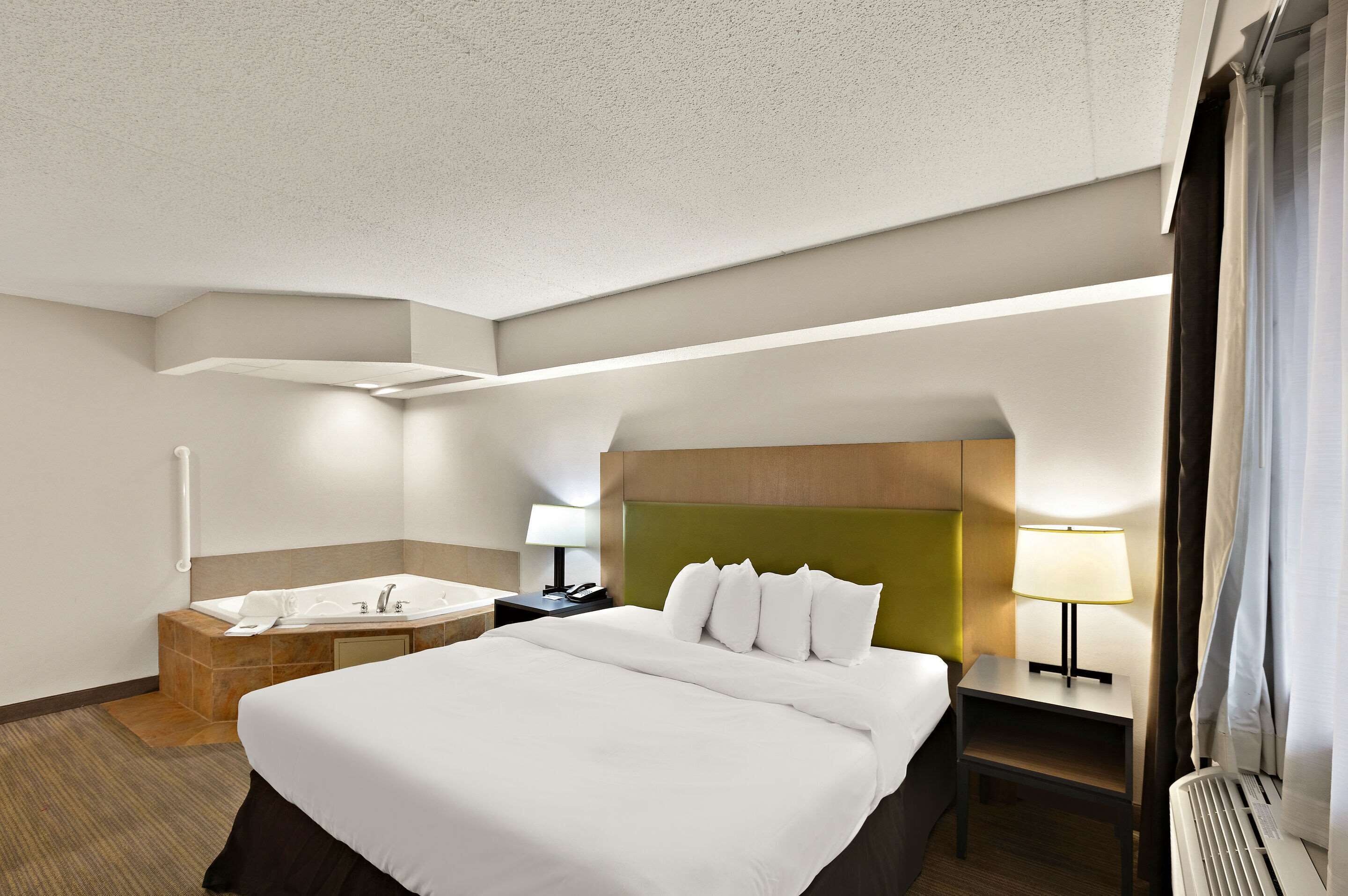 A hotel room with a large bed, a bathroom with a sink and mirror, a nightstand with a lamp, and a window with curtains.