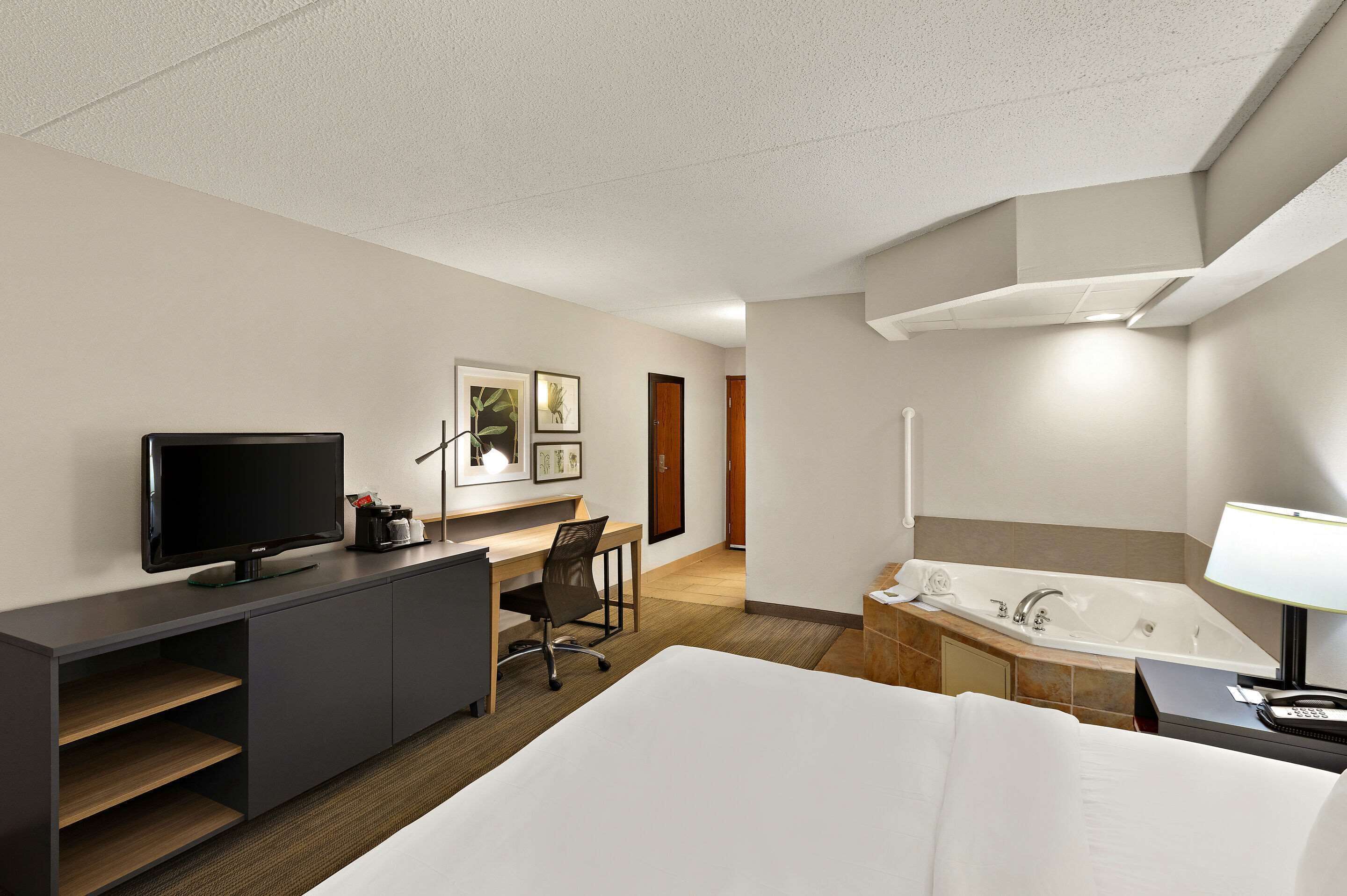 A hotel room with a bed, a desk with a TV, a chair, a bathroom with a bathtub, and a lamp.