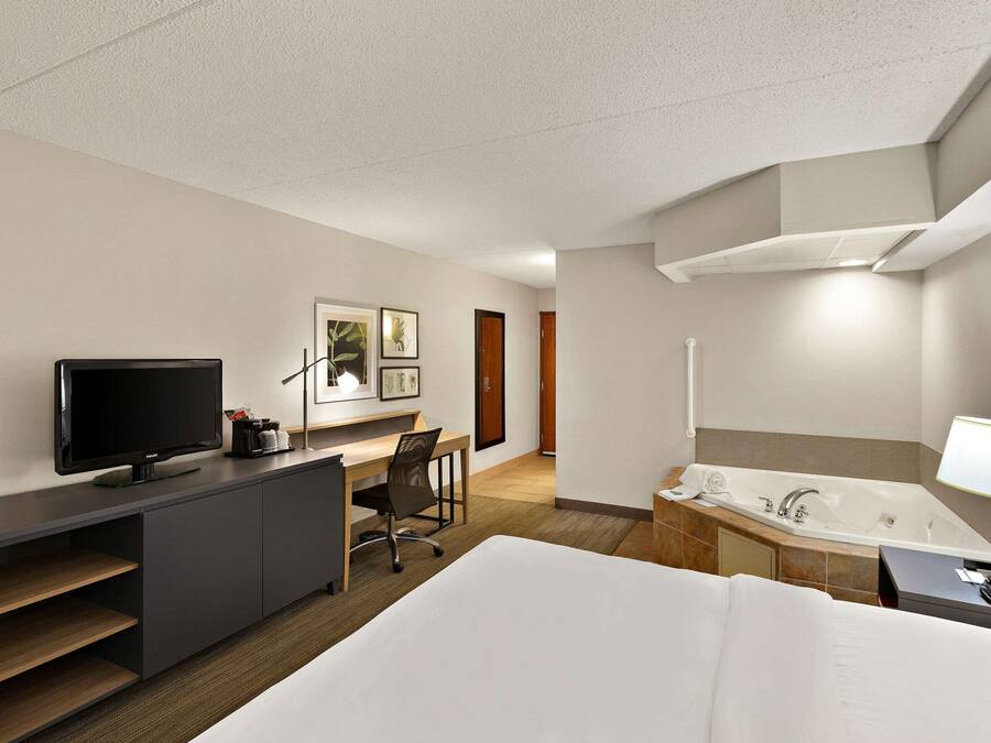 A hotel room with a bed, a desk with a TV, a chair, a bathroom with a bathtub, and a lamp.