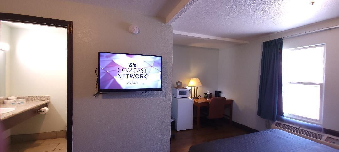 Habitación de hotel con un televisor mostrando el logotipo de Comcast, un microondas, un escritorio con una lámpara y una ventana con cortinas.