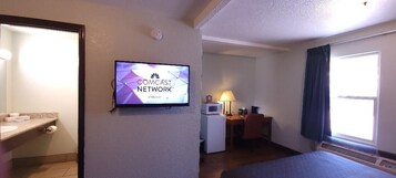 Habitación de hotel con un televisor mostrando el logotipo de Comcast, un microondas, un escritorio con una lámpara y una ventana con cortinas.