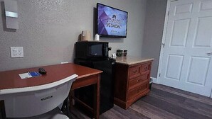 Une petite chambre d’hôtel avec un bureau en bois, une chaise blanche, un four à micro-ondes, un petit réfrigérateur, un téléviseur affichant le logo de Comcast Network et une porte blanche.