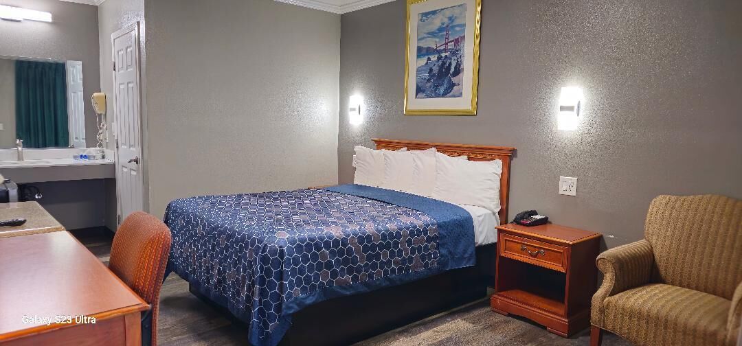 Una habitación de hotel con cama, escritorio, silla, mesita de noche y un cuadro en la pared.