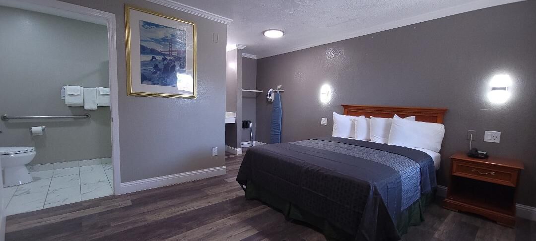 A hotel room with a large bed, a wooden nightstand, a bathroom with a shower, and a painting on the wall.