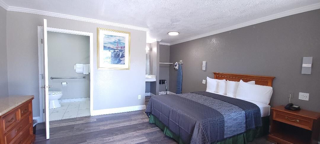 Un dormitorio con cama, una mesita de noche de madera, un aparador y un baño visible a través de una puerta abierta.