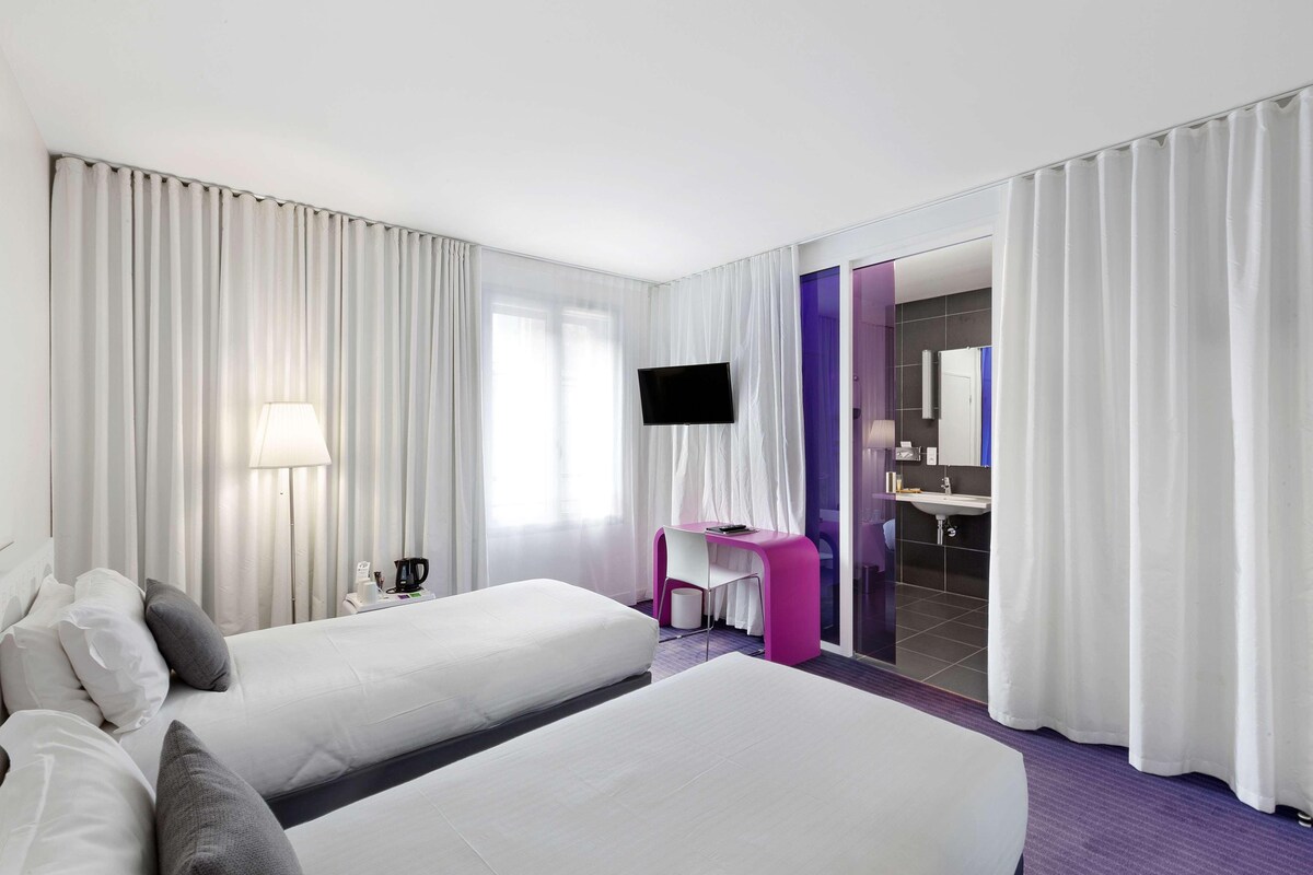 Habitación de hotel con dos camas, un escritorio, televisión y baño visible a través de una puerta de cristal.