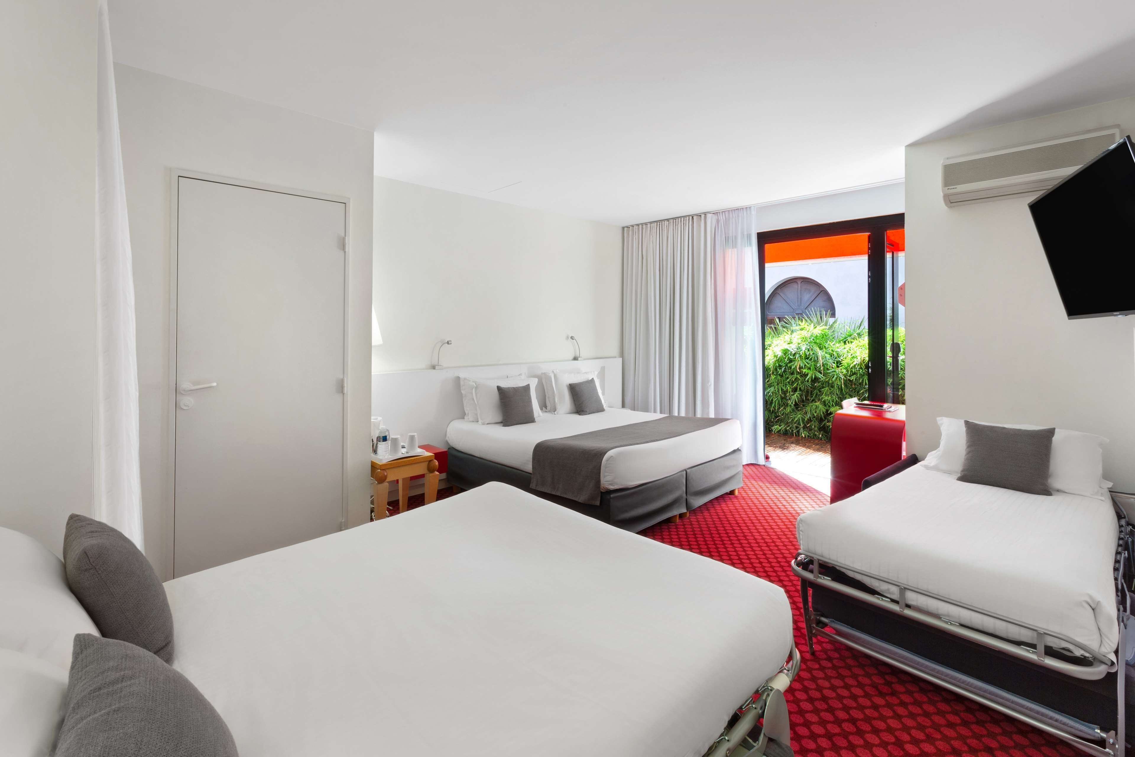 Habitación de hotel con dos camas, alfombra roja, televisión, ventana con cortinas y vista a zonas verdes.