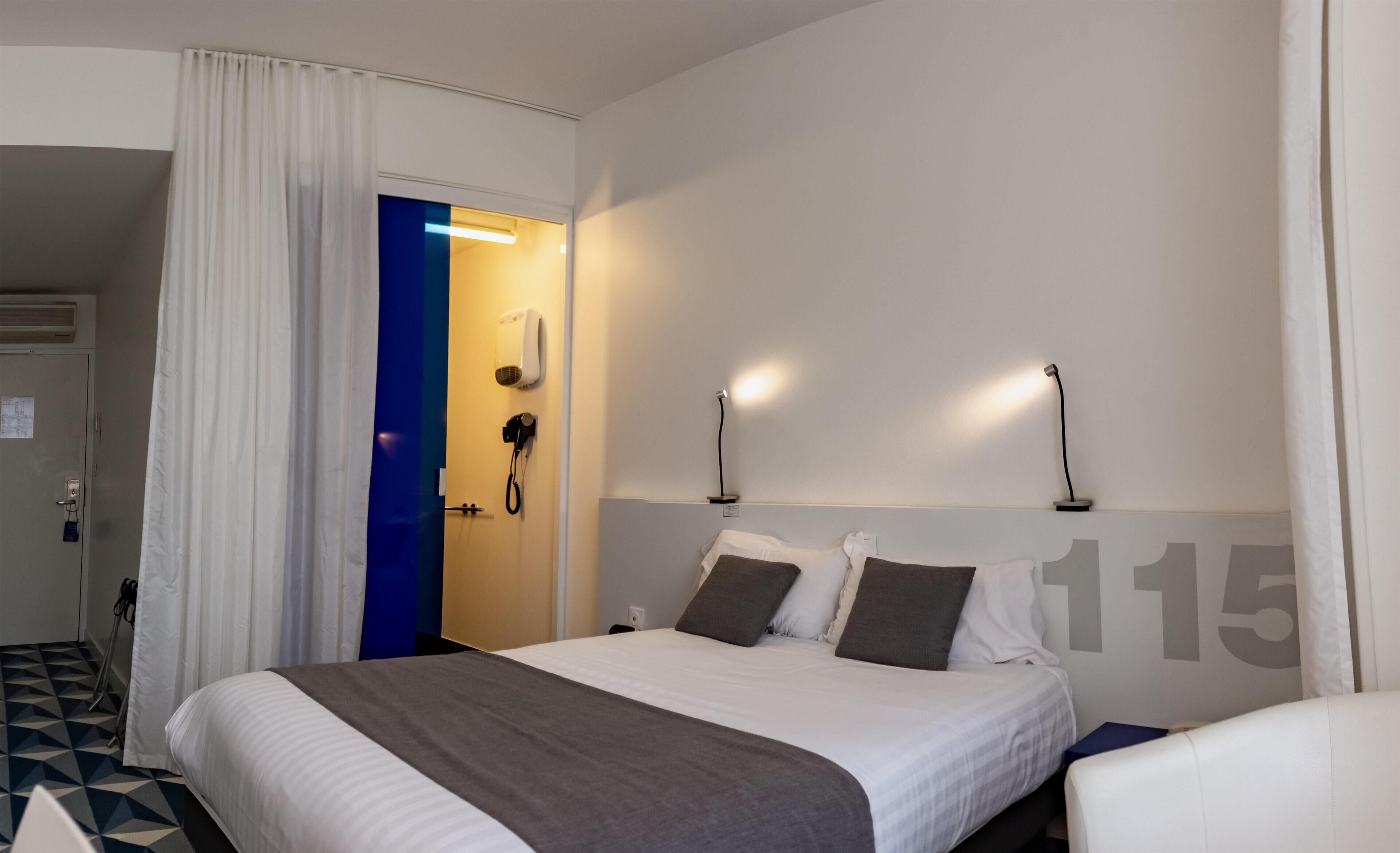 Habitación de hotel con cama, escritorio, silla, baño con ducha y puerta con marco azul.