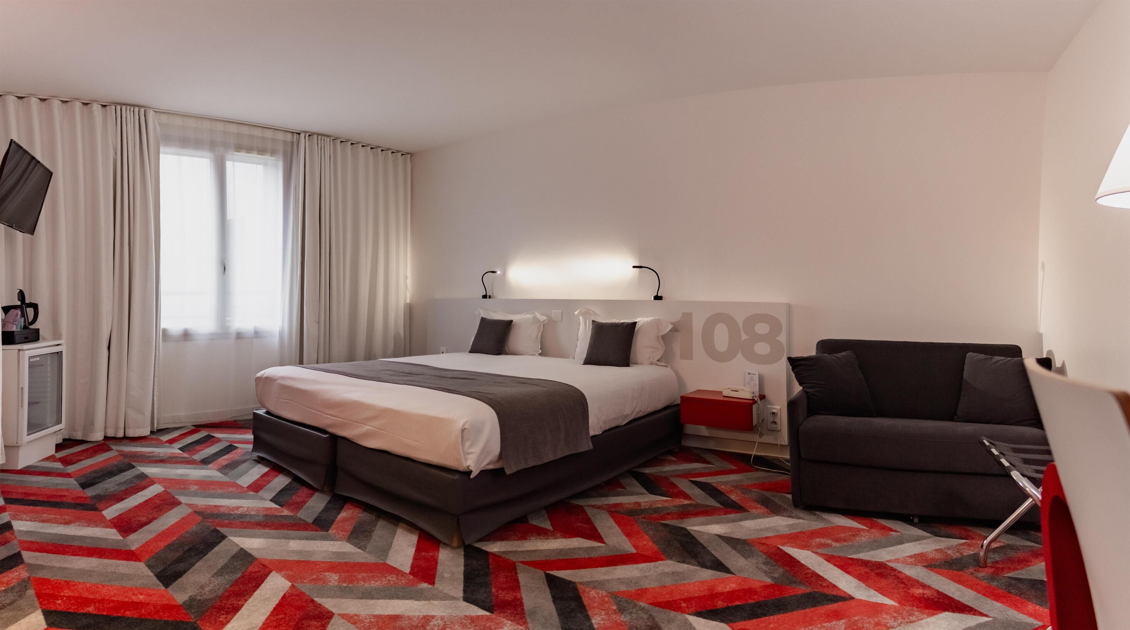A hotel room with a bed, a desk, a chair, and a large window with curtains.