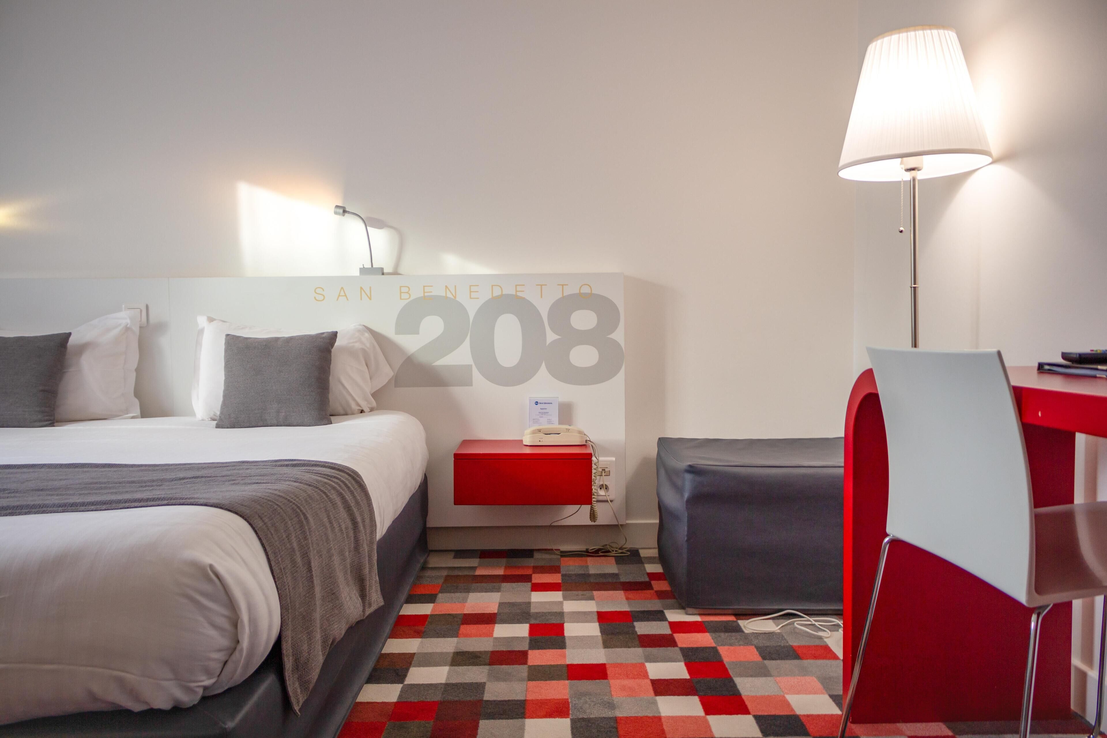 A hotel room with a bed, bedside table, lamp, and a red desk with a chair.