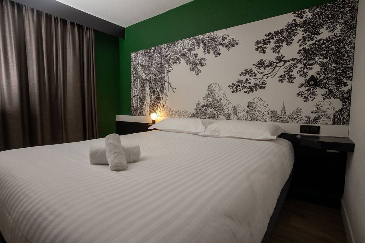 Un lit bien fait, avec du linge de lit et des serviettes blancs, un mur d’accent vert et une fresque murale détaillée en noir et blanc représentant un arbre.