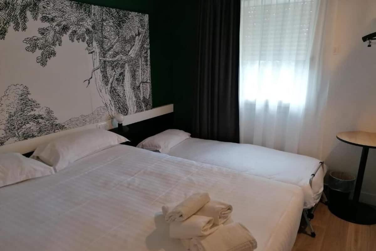 Une chambre d’hôtel avec deux lits, une grande tapisserie murale, une fenêtre avec des rideaux et une table ronde.