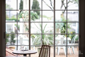 Une vue à travers une fenêtre, avec une table et une chaise, un verre d’eau, et un espace extérieur aménagé avec des chaises et des plantes.