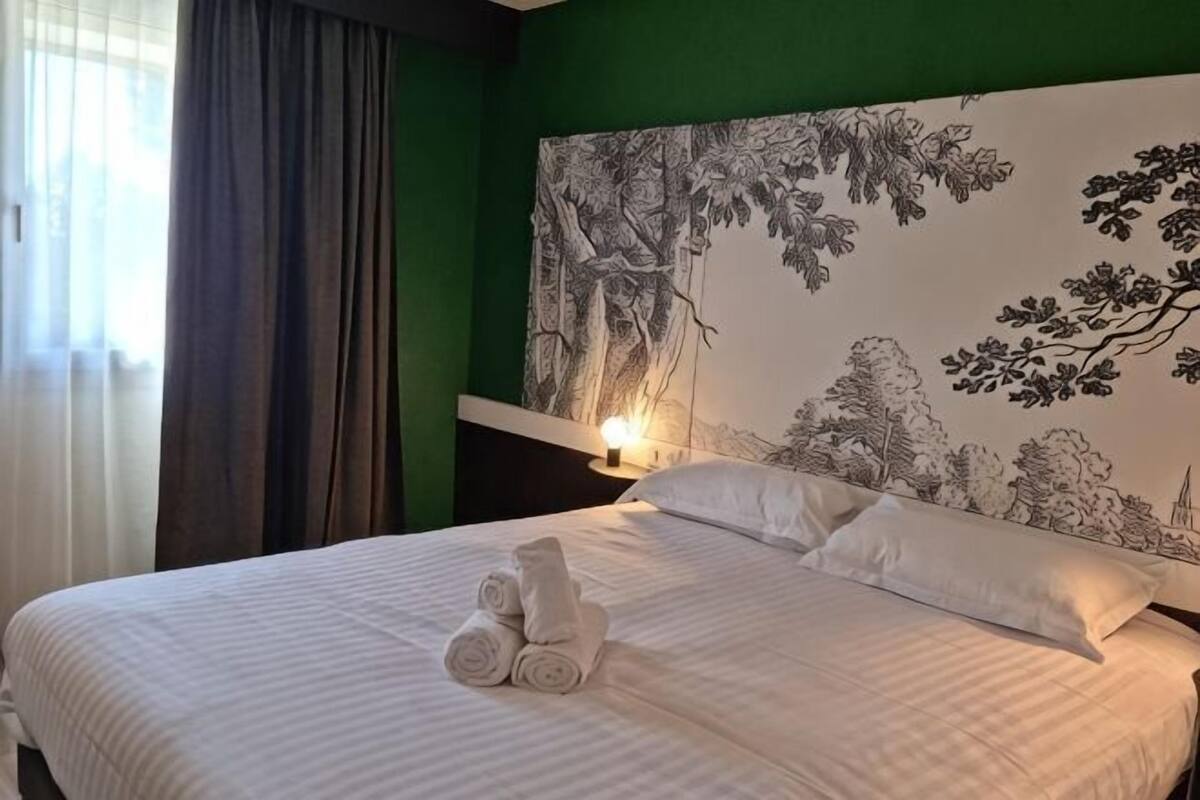 Un lit bien fait, avec des serviettes pliées, une tête de lit ornée d’un motif représentant un arbre et ses branches, et une lampe de chevet.