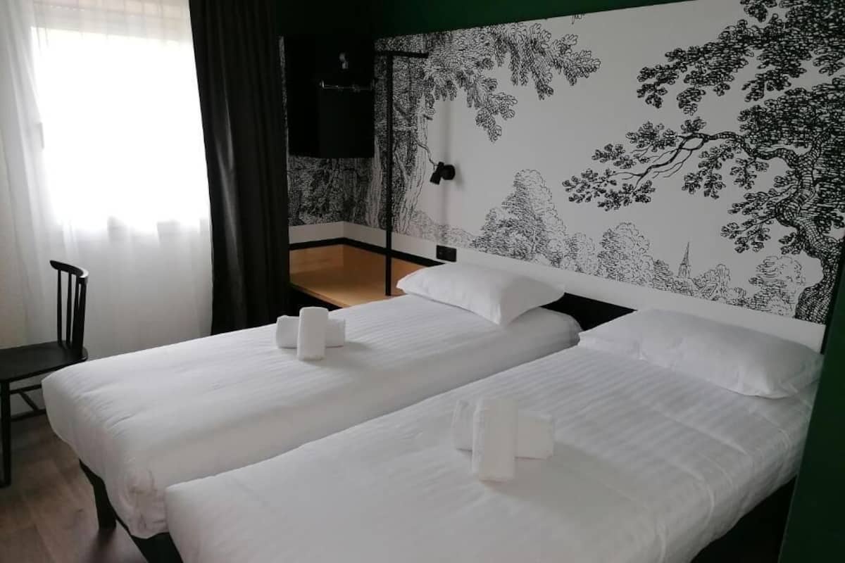 Une chambre d’hôtel avec deux lits, une grande fenêtre avec des rideaux légers, un mur orné d’un motif d’arbre en noir et blanc et un plancher en bois.