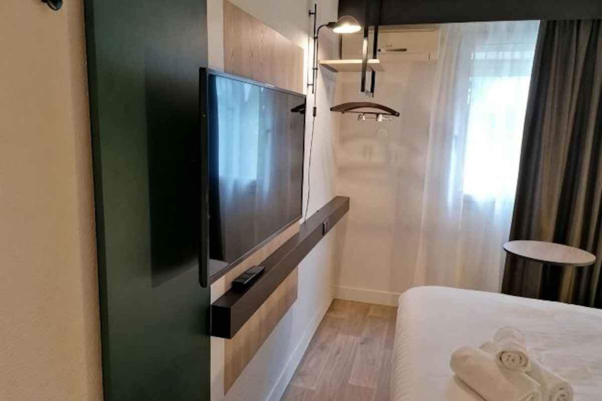 A hotel room with a flat-screen TV mounted on the wall, a bed with neatly folded towels, a small round table, and a window with curtains.