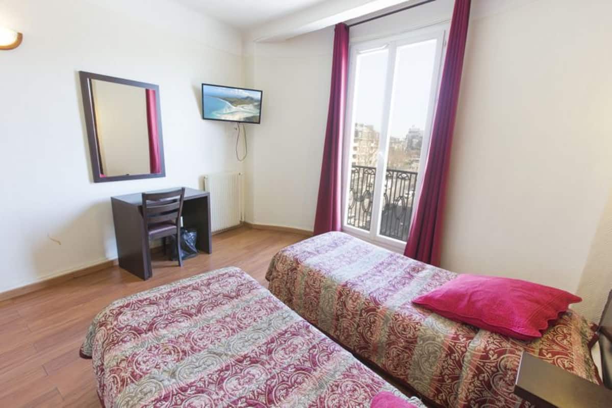 Habitación de hotel con dos camas, un escritorio, una silla, un televisor y ventana con vistas a edificios.