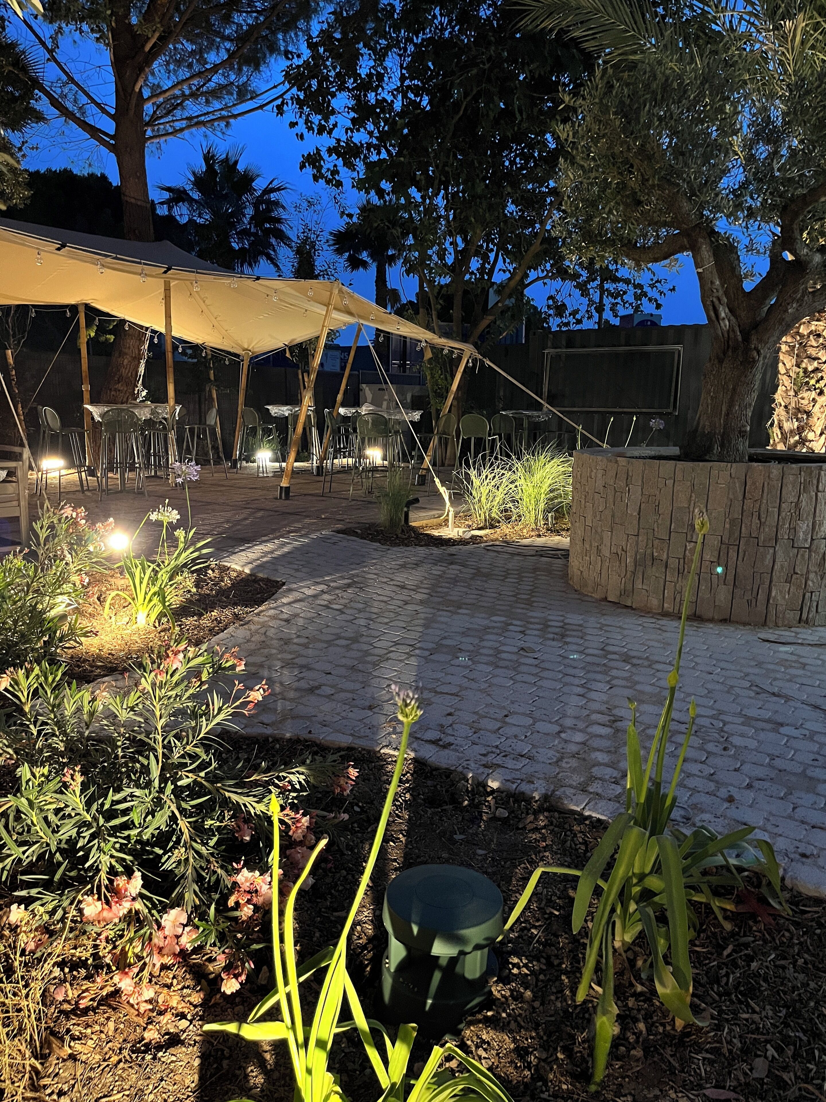 An outdoor patio area with tables and chairs under a canopy, surrounded by plants and a stone wall.