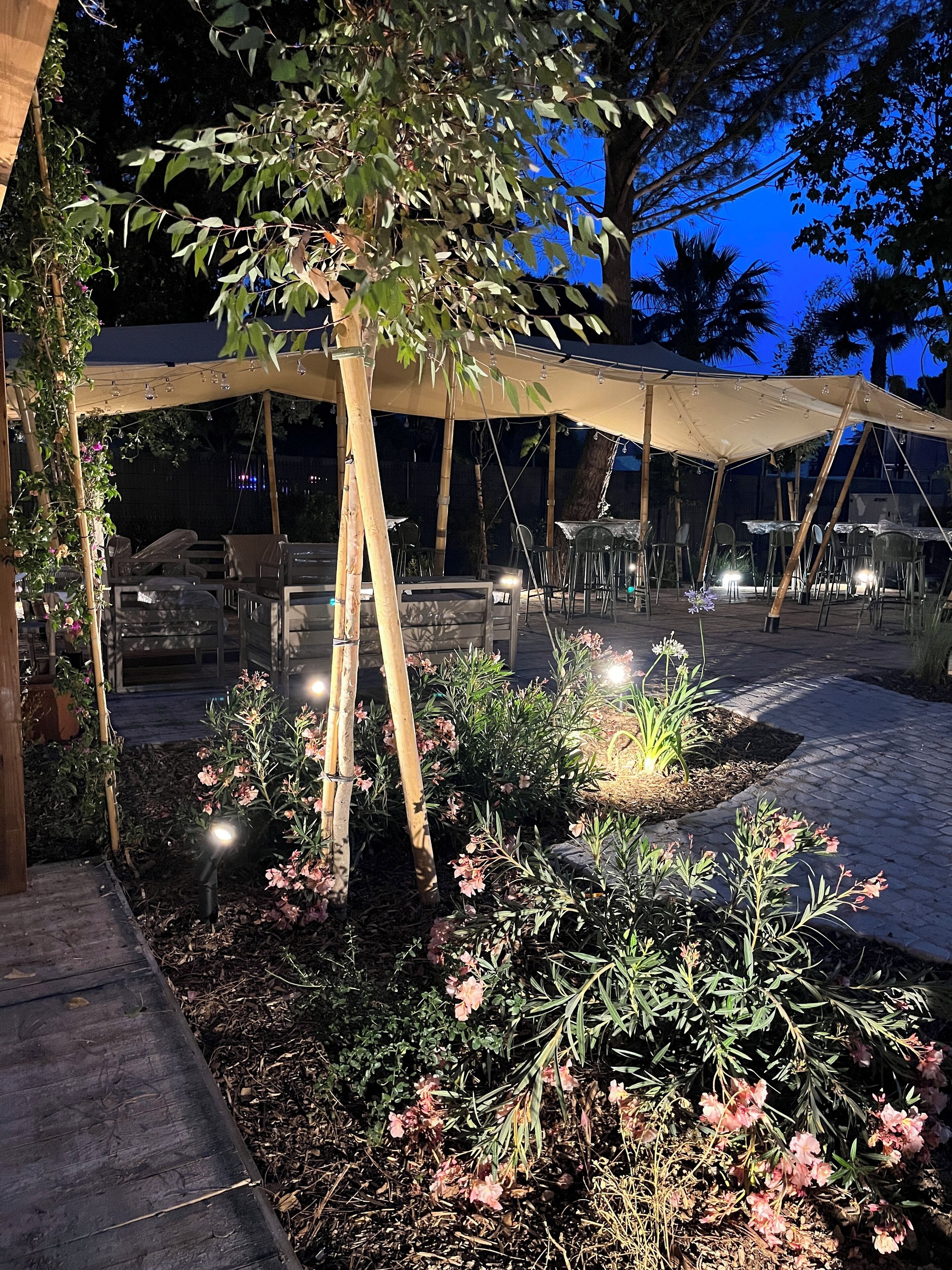 Un área de estar al aire libre con mesas y sillas bajo una pérgola, rodeada de plantas y flores.