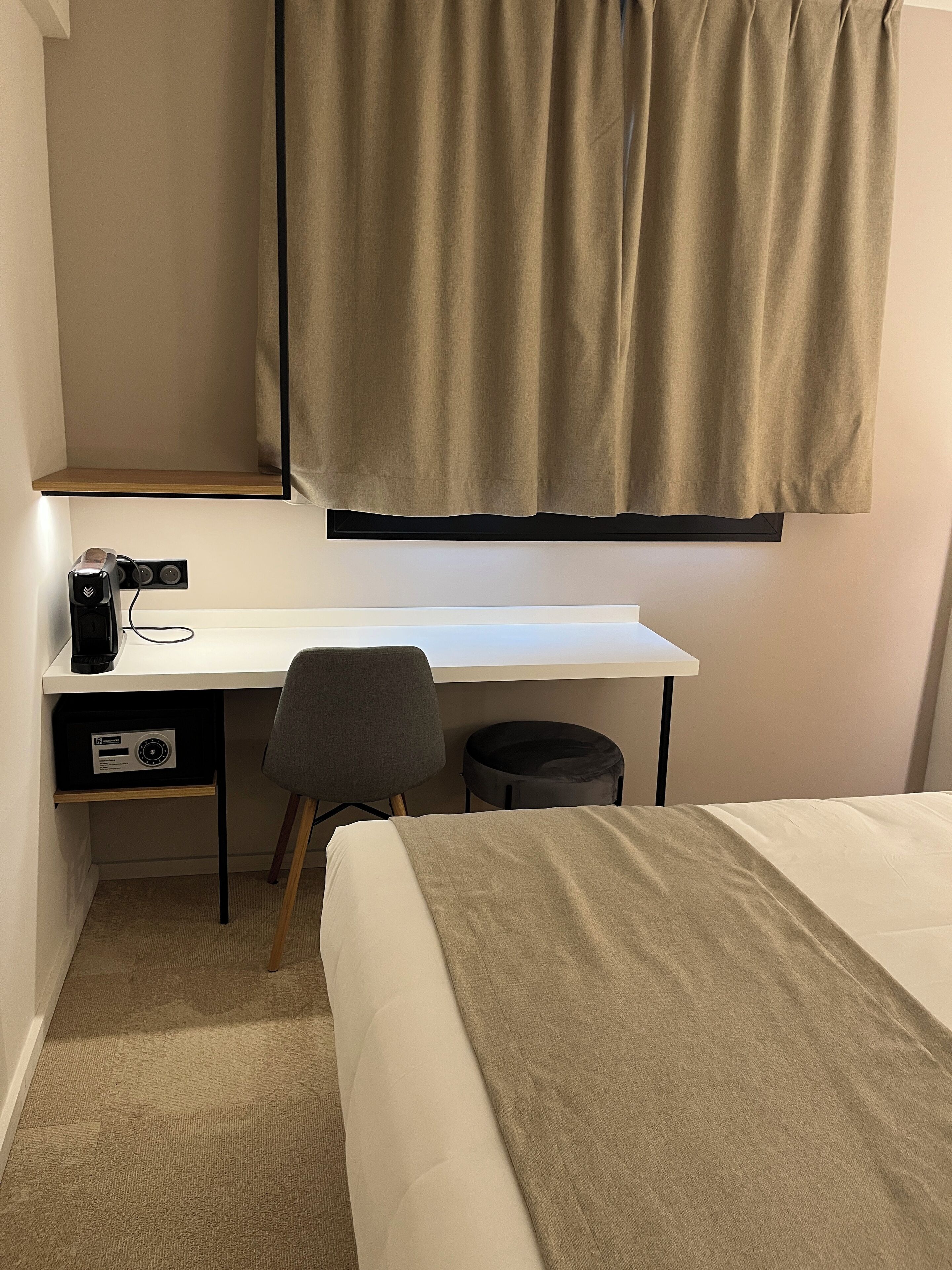 Quarto de hotel compacto com uma cama de solteiro, uma mesa com cadeira, um banquinho preto pequeno e uma janela com cortinas.