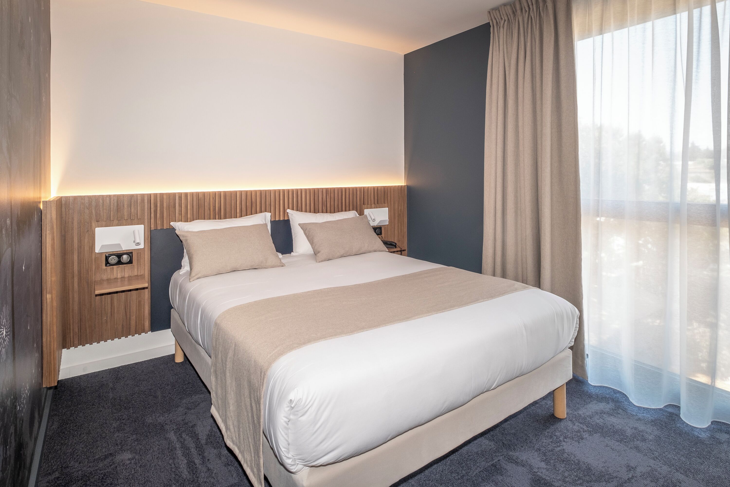 Una habitación de hotel moderna con una cama grande, una mesita de noche con lámpara y vistas al exterior a través de cortinas translúcidas.