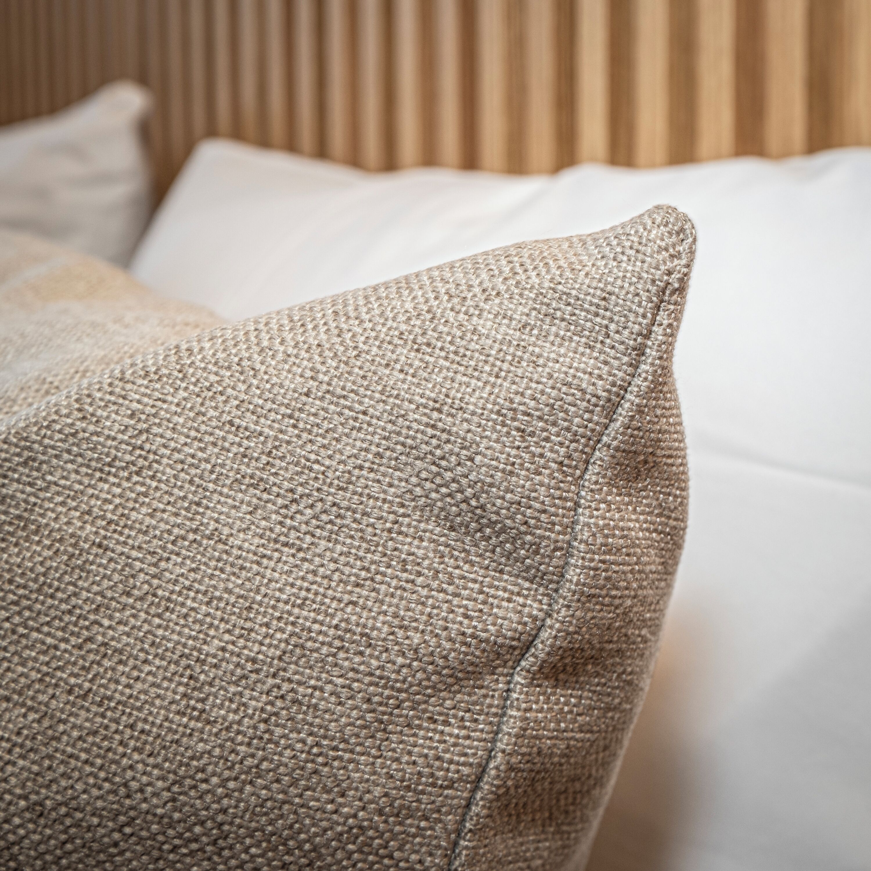 Gros plan d’un coussin beige à texture particulière, posé sur un lit recouvert de draps blancs.