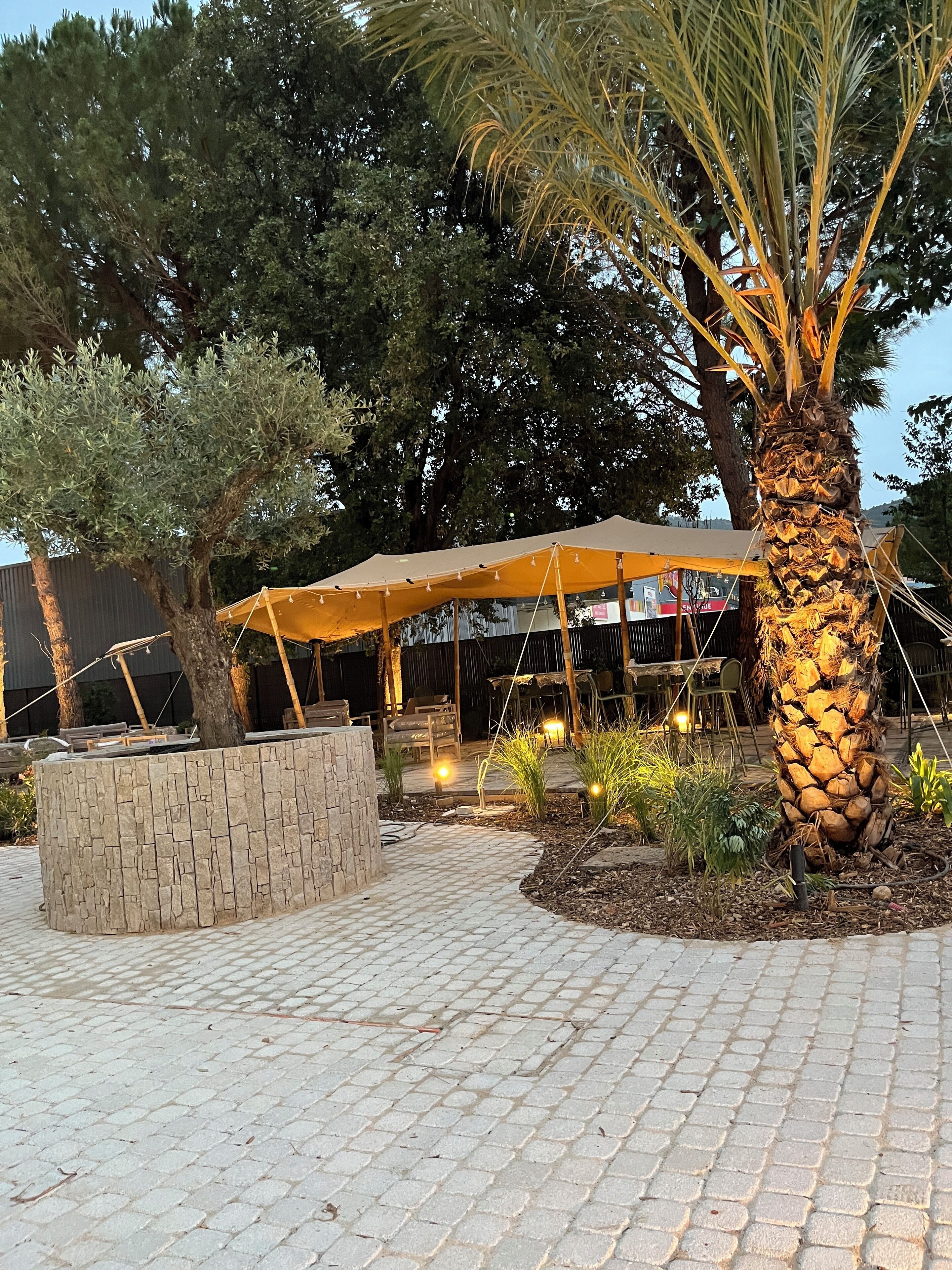 An outdoor patio area with a stone planter, a palm tree, and a covered seating area with string lights.