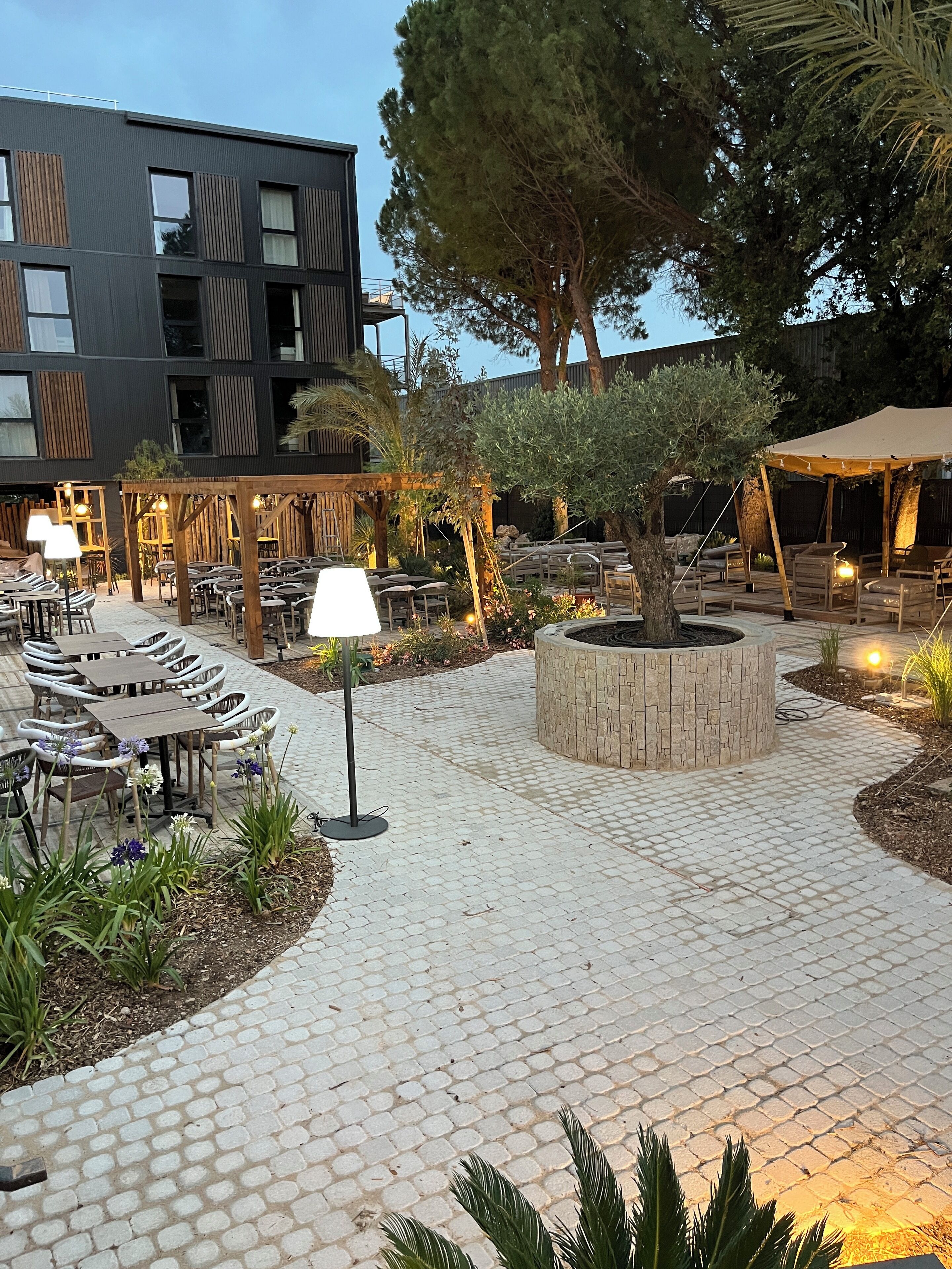 An outdoor patio area with tables and chairs, a stone fire pit, and a pergola with string lights.
