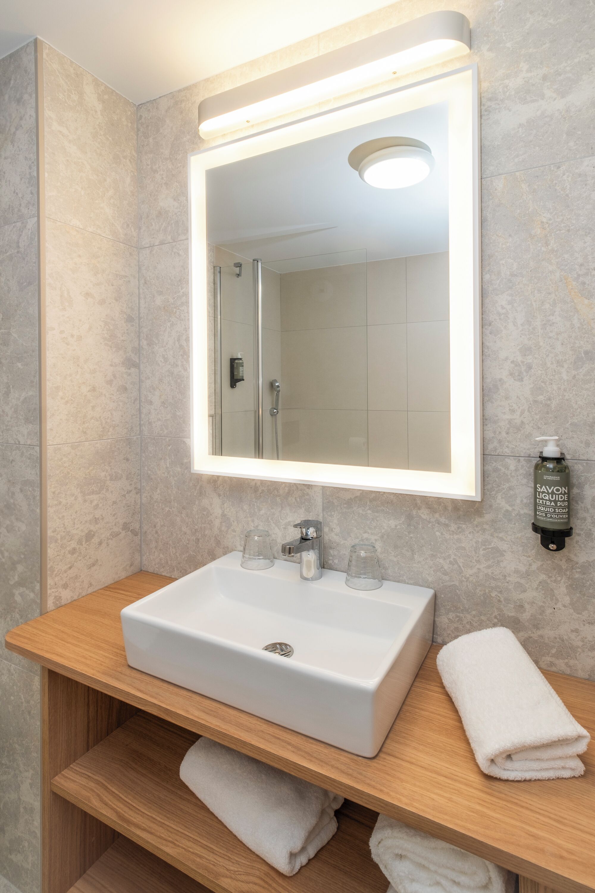 Un baño con lavamanos blanco, un espejo con iluminación integrada, un perchero y una botella de jabón.