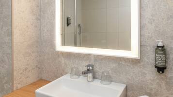 Un baño con lavamanos blanco, un espejo con iluminación integrada, un perchero y una botella de jabón.