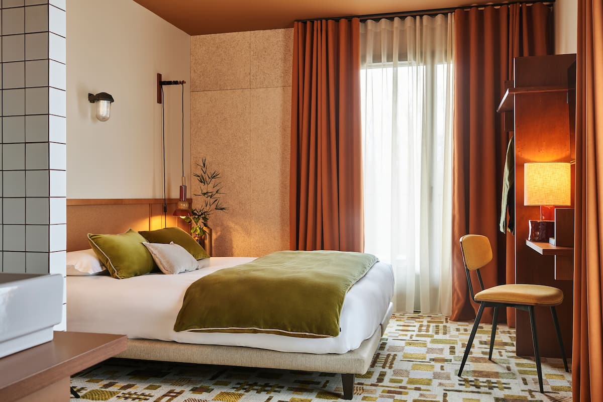 Habitació d'hotel amb un llit, una cadira, una làmpada i una finestra amb cortines.