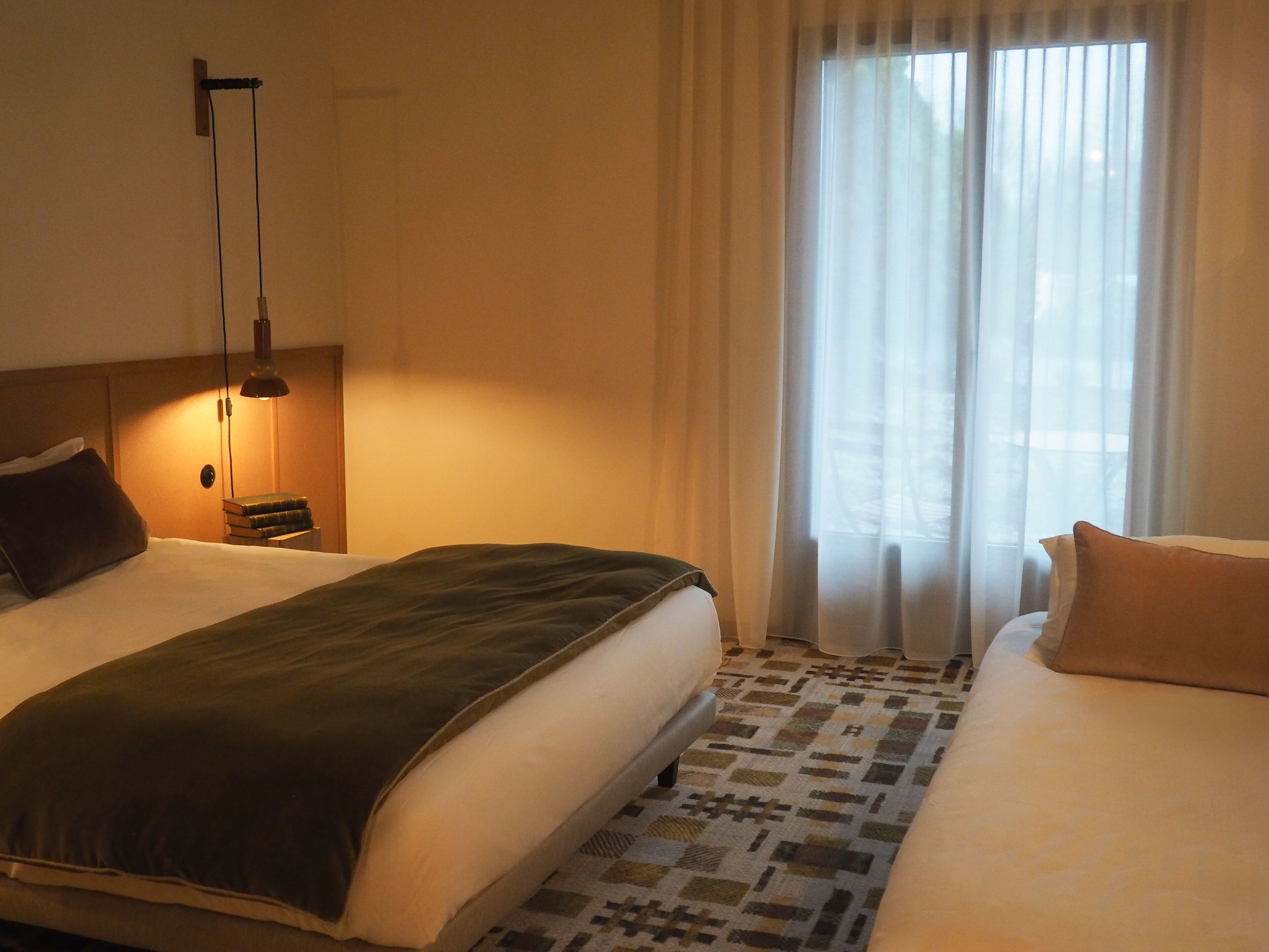 A hotel room with a large bed, a smaller bed, a nightstand with a lamp, and a window with sheer curtains.