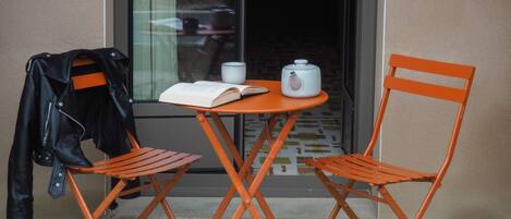 A small table with two orange chairs, a black leather jacket hanging on the wall, and a book and teapot on the table.