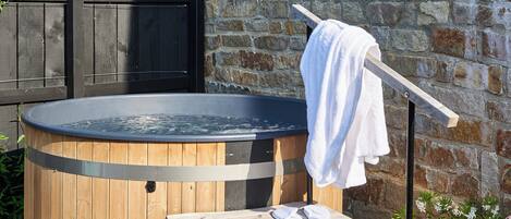 A wooden hot tub with a towel on a rack, set against a stone wall with a wooden fence in the background.