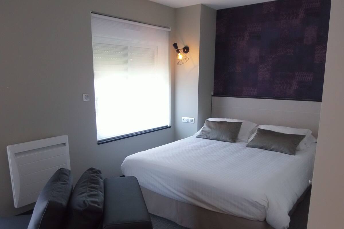 A bedroom with a bed, a window with blinds, a wall-mounted light, and a radiator.