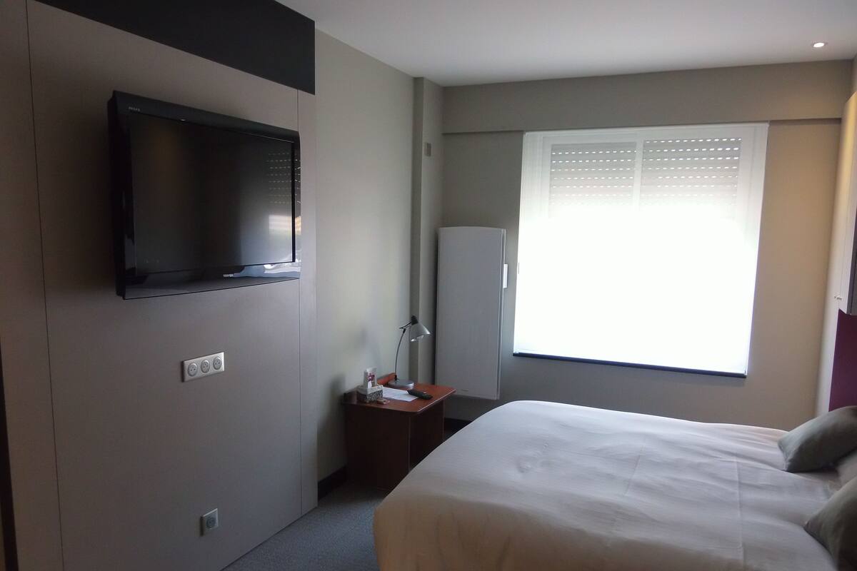 A hotel room with a bed, a TV mounted on the wall, a desk with a lamp, and a window with blinds.