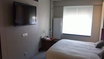 Una habitación de hotel con cama, televisión montada en la pared, escritorio con lámpara y ventana con persianas.