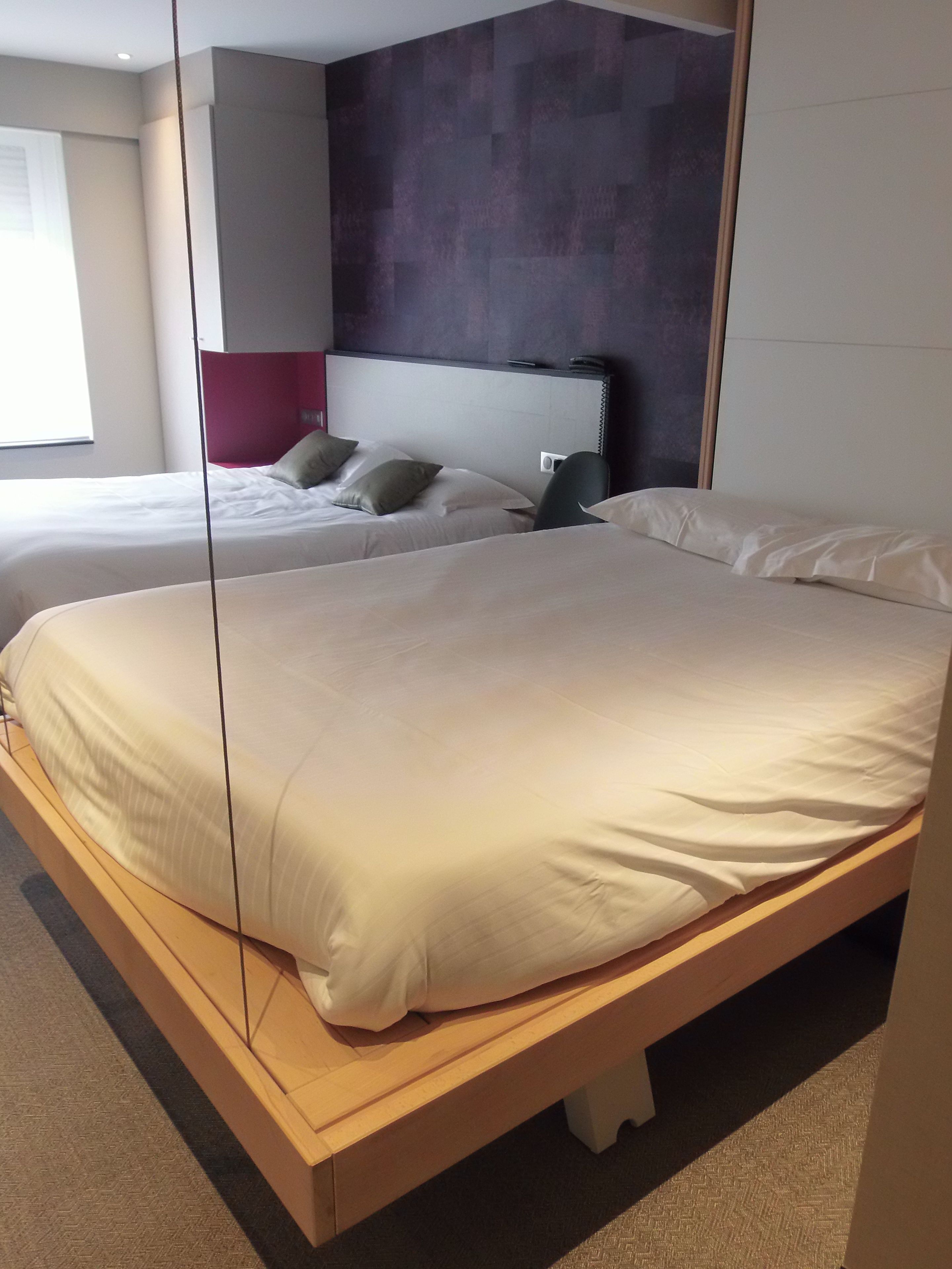 Habitació d'hotel amb dos llits, un panell de capçal modern i un mirall gran.