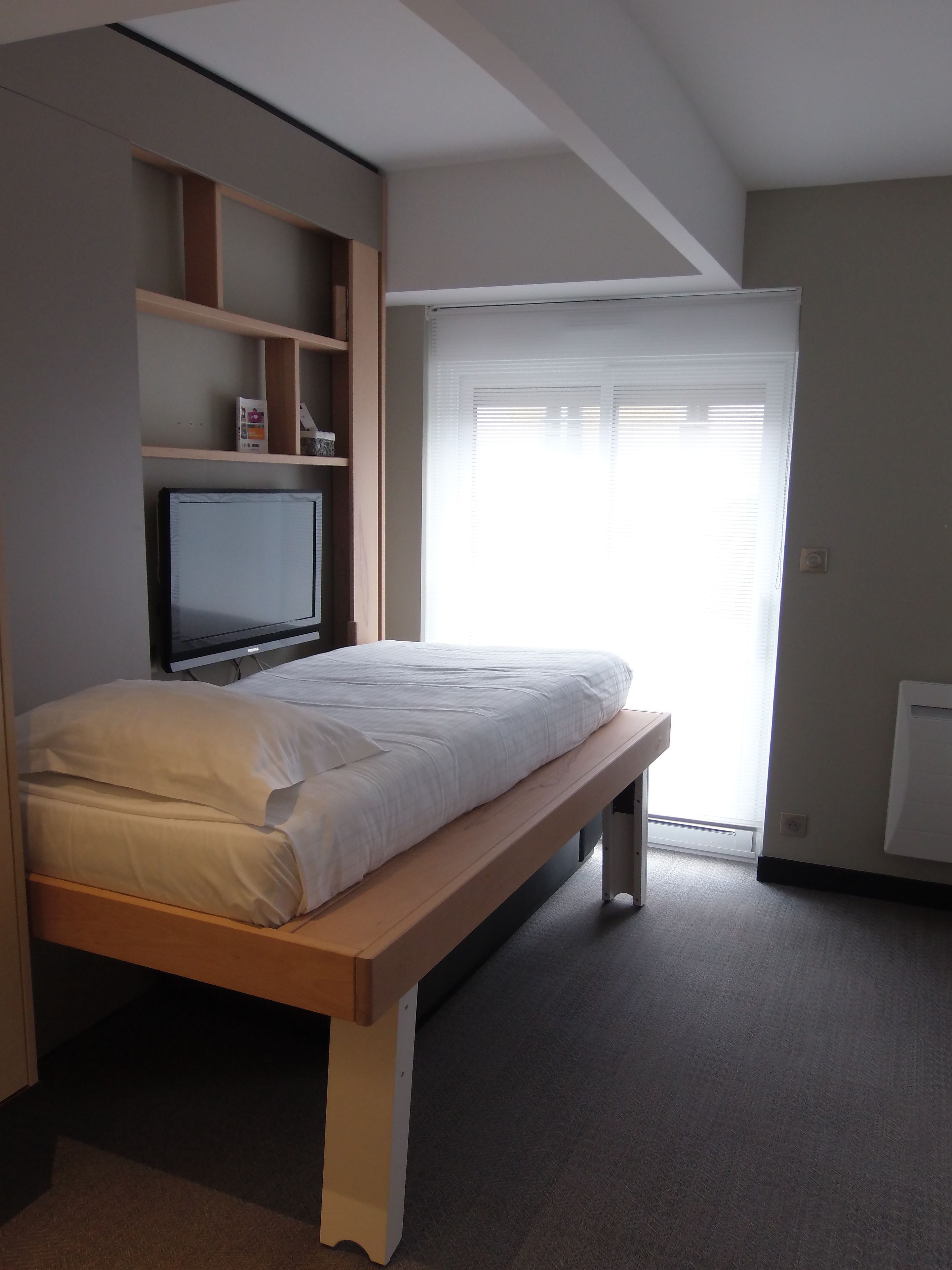 A bedroom with a bed, a TV mounted on the wall, and a window with blinds.