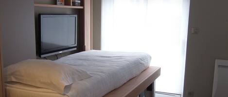 Un dormitorio con cama, televisor montado en la pared y ventana con persianas.