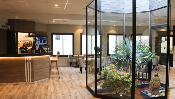 A modern bar area with a wooden floor, a glass enclosure with a plant and decorative items, and a television screen. - The Originals Boutique, Hotel La Chaussairie, Rennes (Chartres-de-Bretagne)